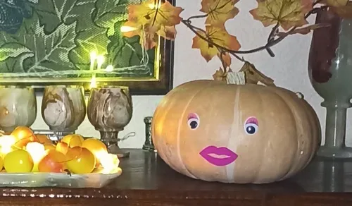 A light brown pumpkin sits on a dark wooden table, decorated with cartoonish pink eyes and bright pink lips, giving it a…