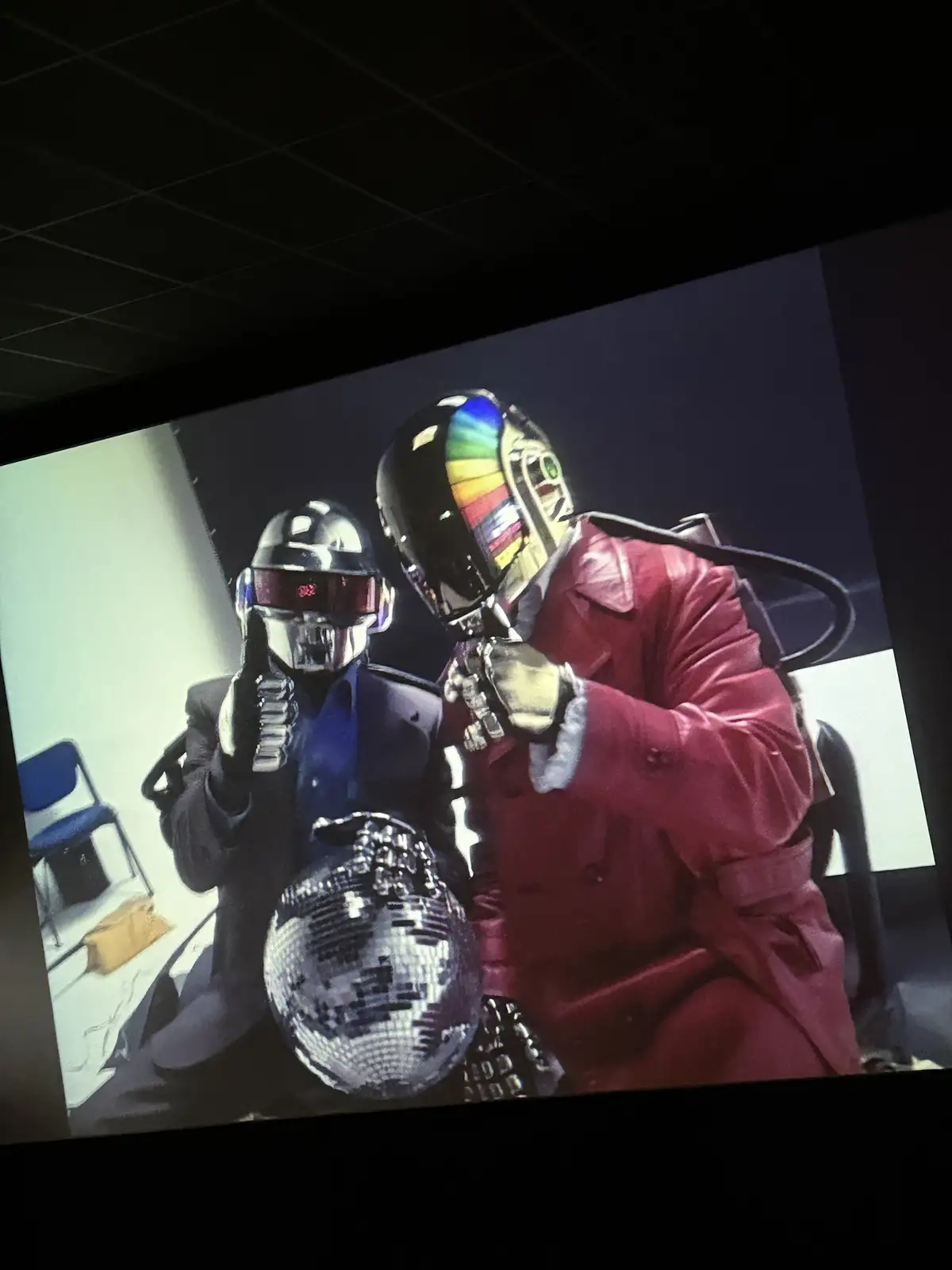 A photo of the electronic music duo Daft Punk, shown on a screen, with one member in a black suit and helmet and the other…