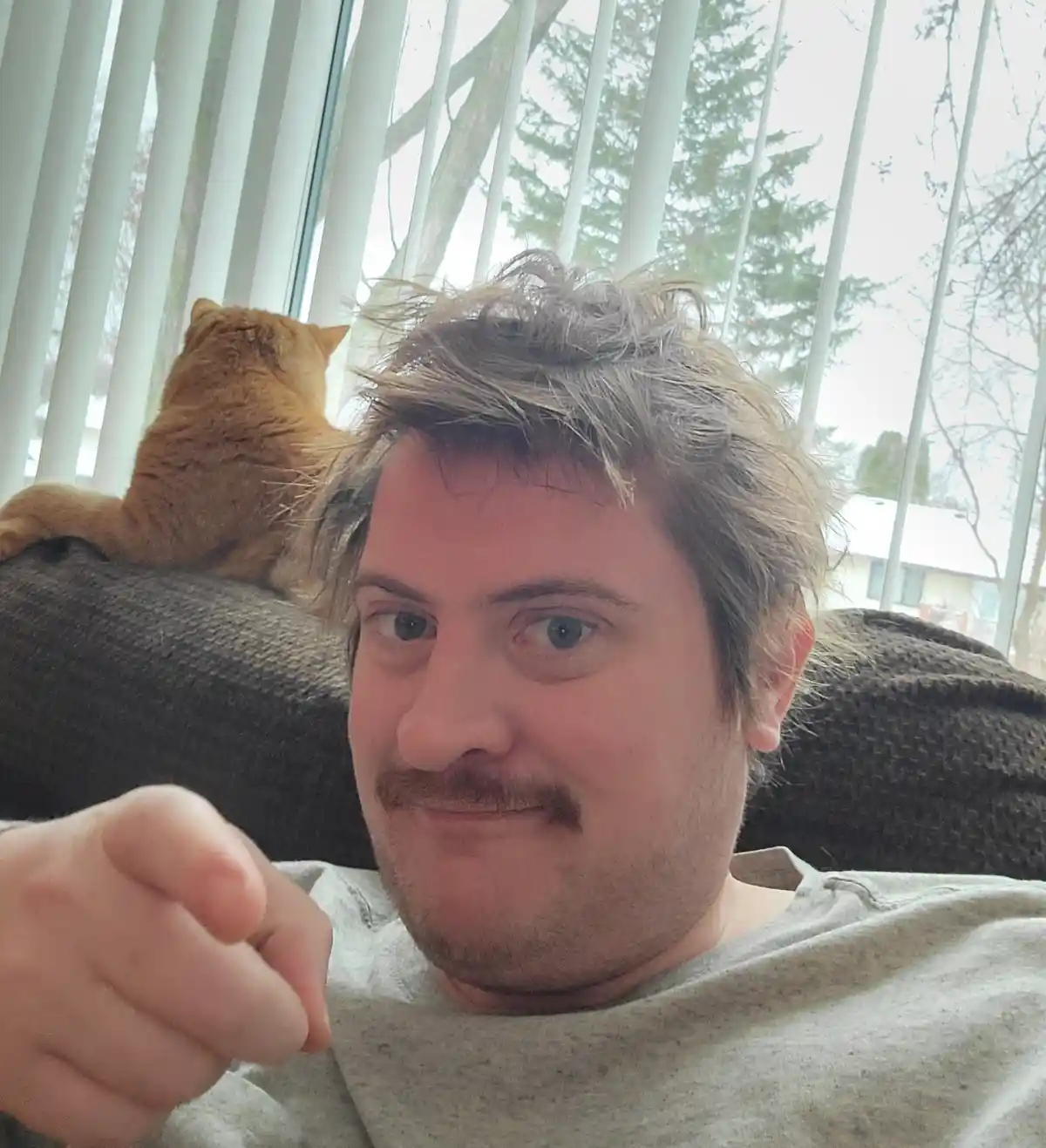A man with messy hair and a mustache sits on a couch, pointing at the camera with a smug expression, while a ginger cat…