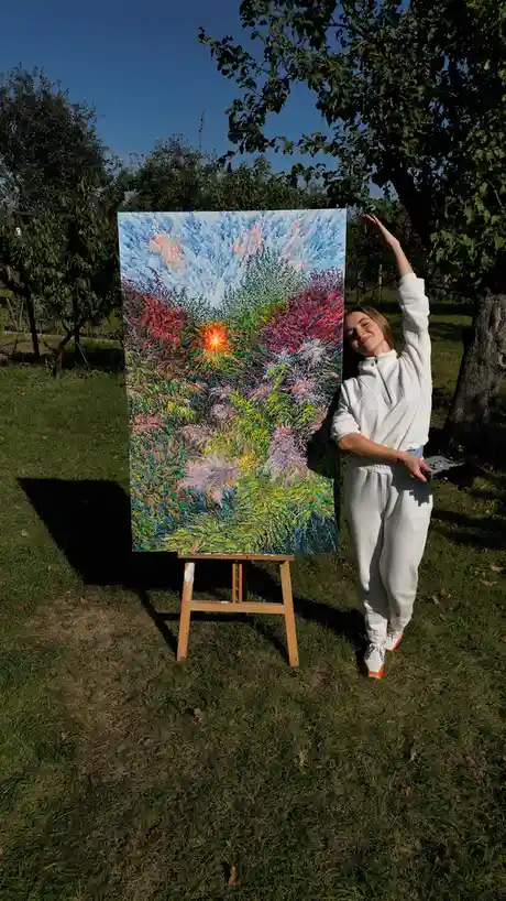 A woman in a white sweatsuit stands outdoors next to a large, colorful abstract painting on an easel. She holds a drone…
