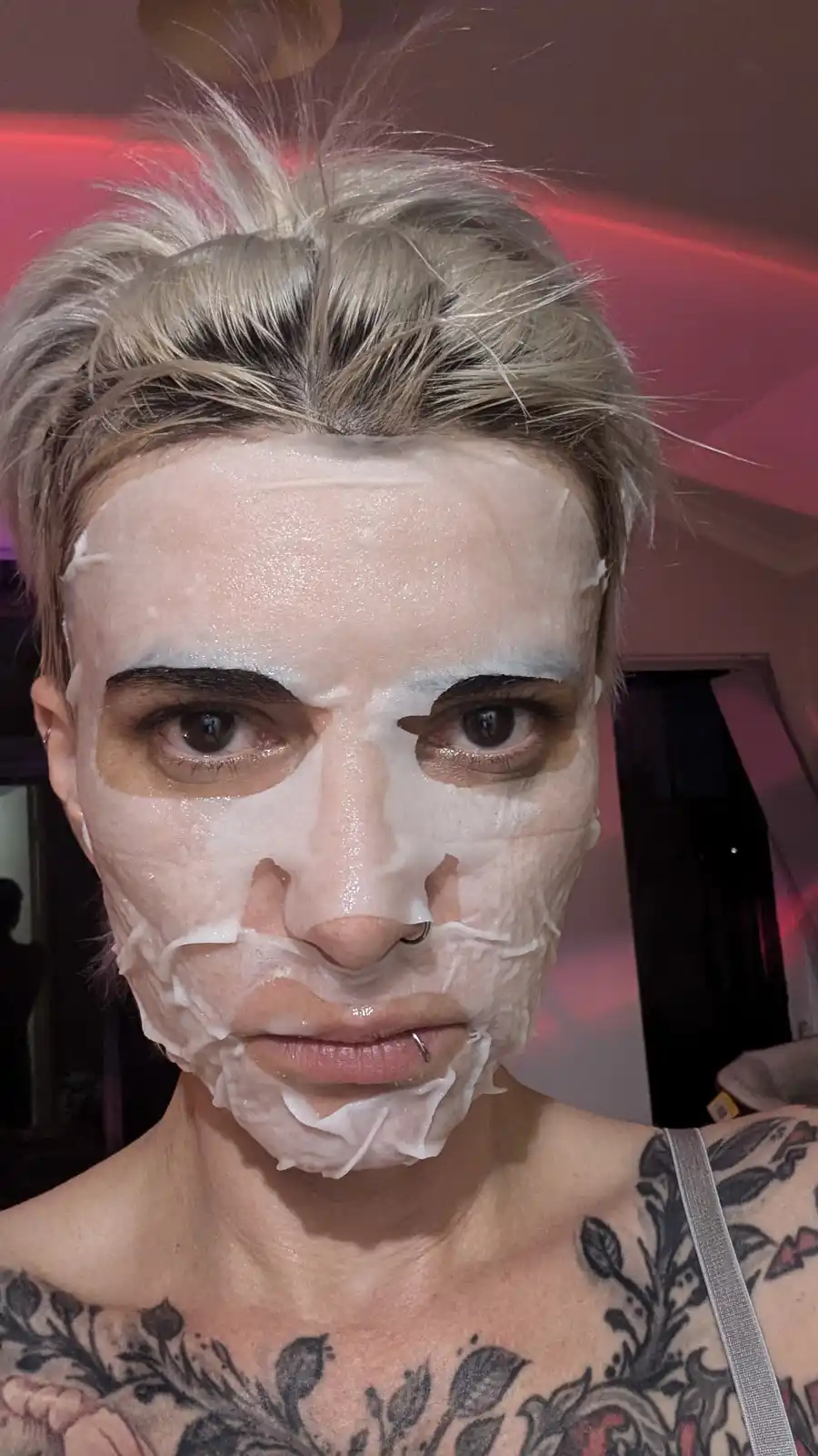 A person with short blonde hair and visible tattoos on their shoulders has a white sheet mask applied to their face, with…