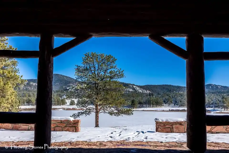 A scenic winter photo taken from inside a wooden structure, looking out over a snow-covered landscape with a lone tree in…