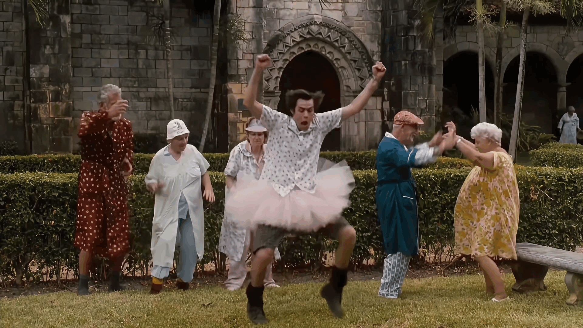The image shows a man in a white shirt and pink tutu jumping energetically in a garden while surrounded by other people in…