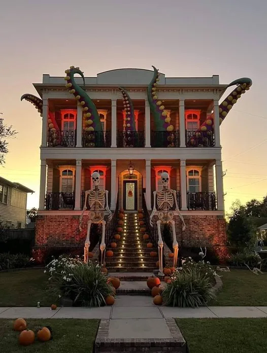 a two-story house decorated for halloween with two large skeleton figures standing at the entrance, glowing pumpkins lining…