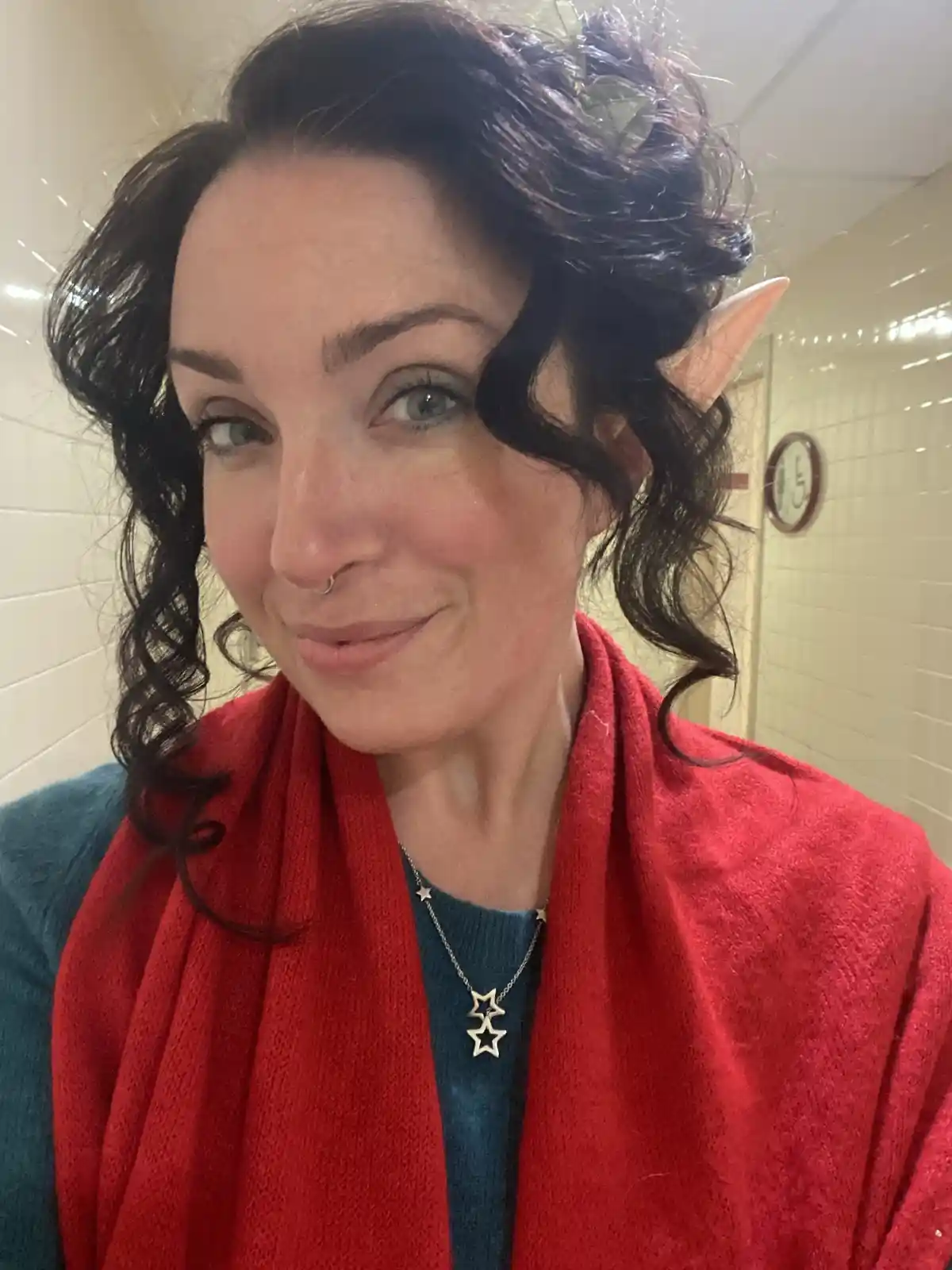 A woman with dark curly hair, wearing a red scarf and a blue sweater, smiles at the camera while sporting a single pointed…
