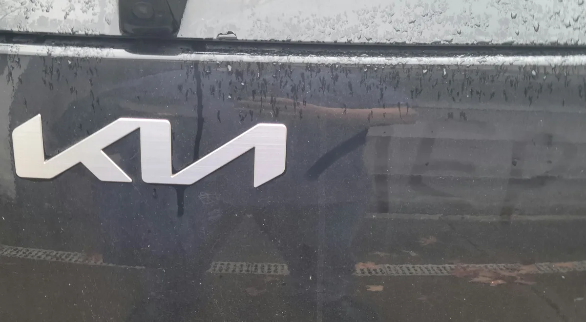 A close-up of a wet car door featuring a silver Kia logo, with raindrops covering the surface and a faint reflection of a…