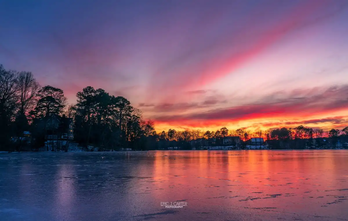 A serene winter sunset over a frozen lake, with vibrant pink, orange, and purple clouds reflecting on the icy surface…