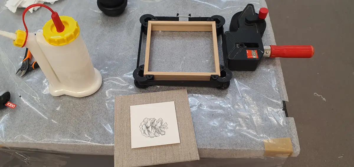 A workbench holds a small pinecone drawing on paper, a wooden picture frame held in a black clamp, a glue bottle, and a…