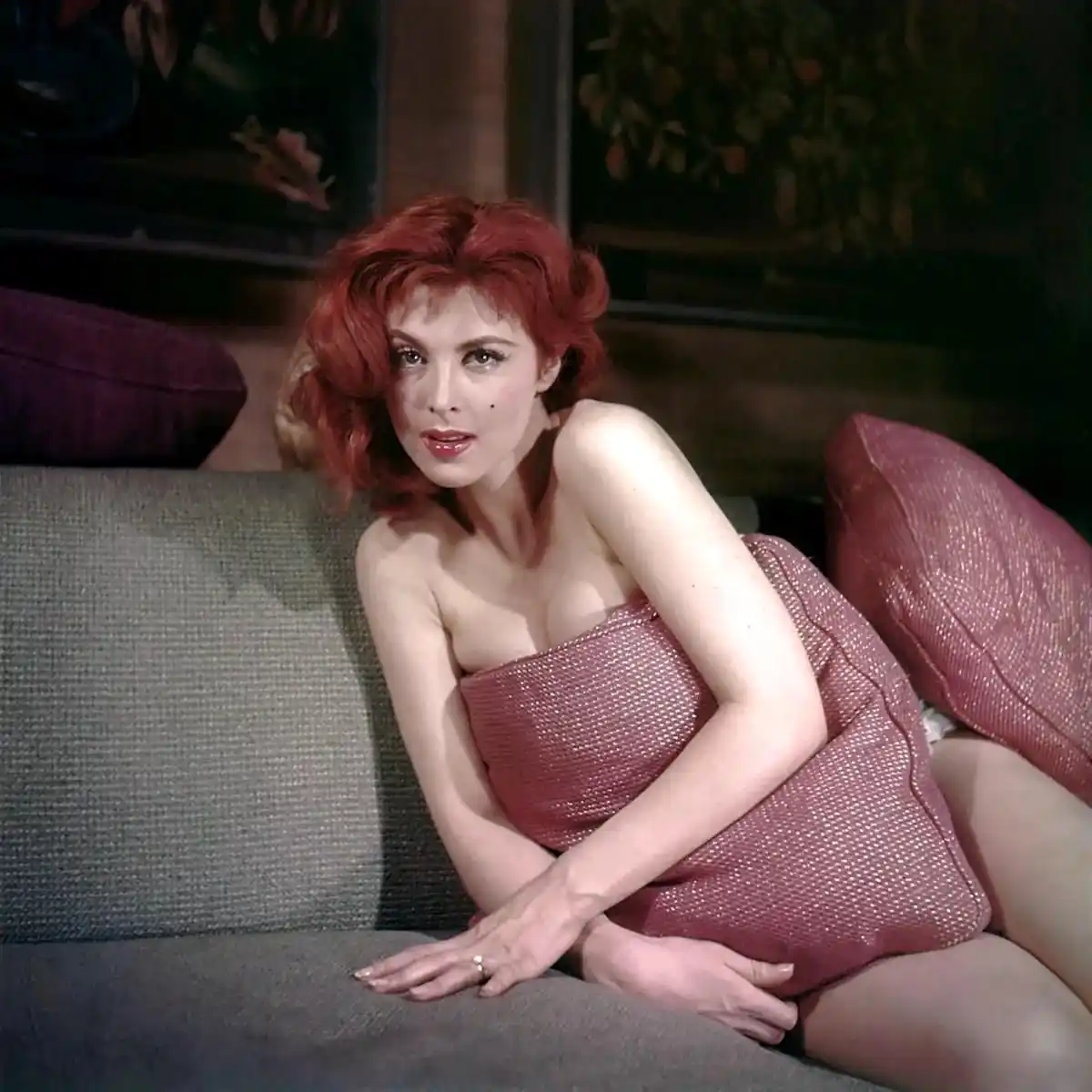 a woman with red hair and a sparkly pink strapless dress sits provocatively on a gray couch, looking directly at the camera…