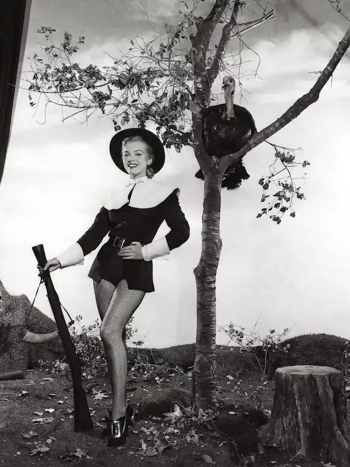 A vintage black-and-white photo shows a smiling woman in a colonial-style outfit with a hat, holding a rifle, standing next…