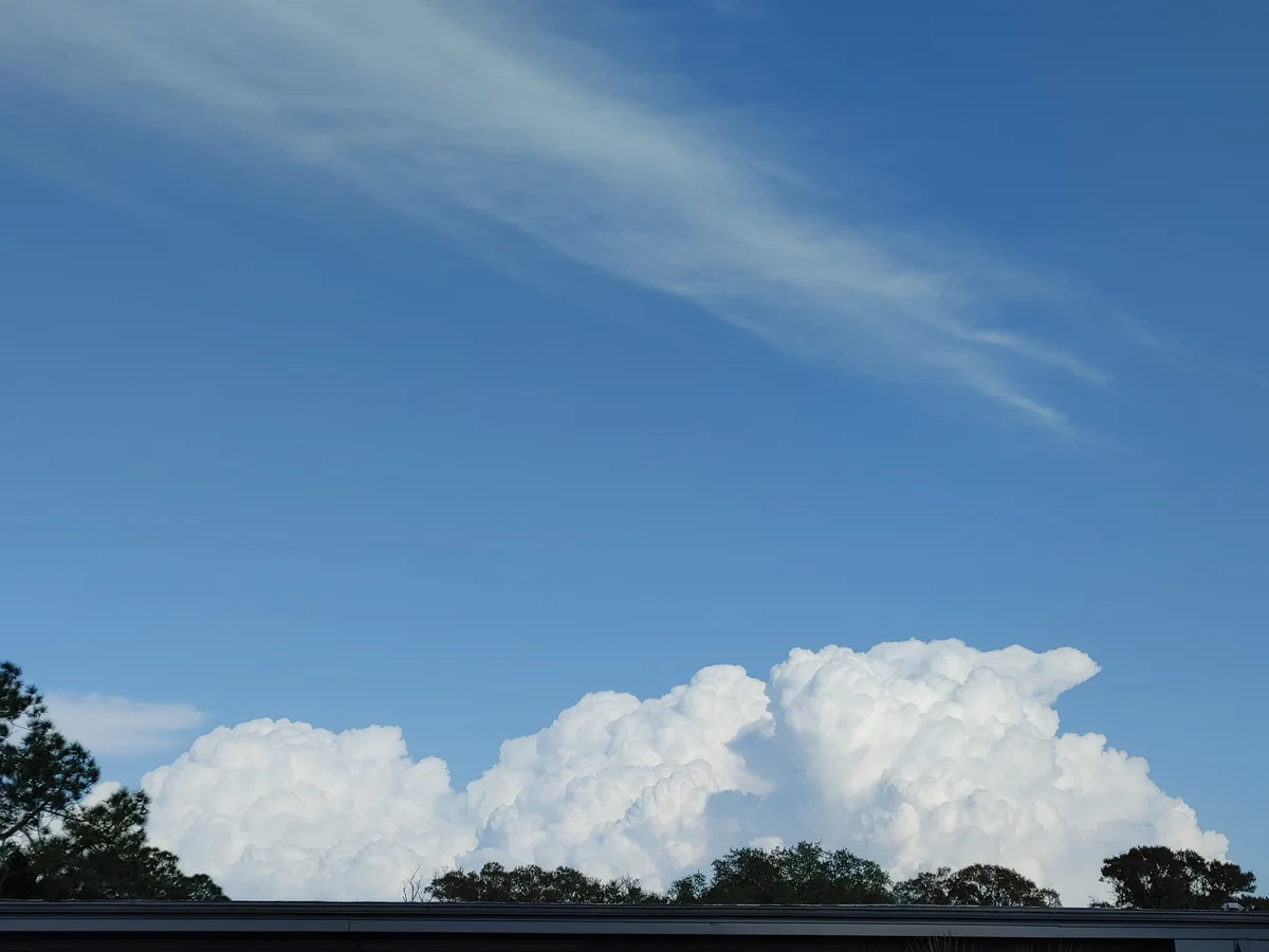 A photo of a large, fluffy white cloud formation against a clear blue sky, with tree tops and a roofline visible at the…