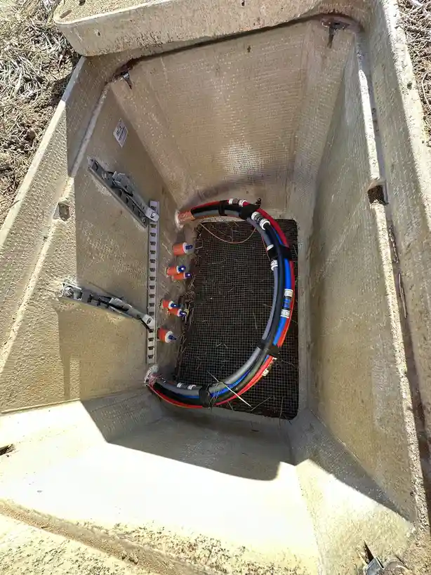 An overhead view of an open concrete utility box containing a large, curved bundle of red, blue, and gray hoses secured with…