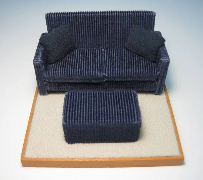 A miniature, dark blue corduroy sofa with two matching pillows and a matching ottoman, all displayed on a beige carpeted…