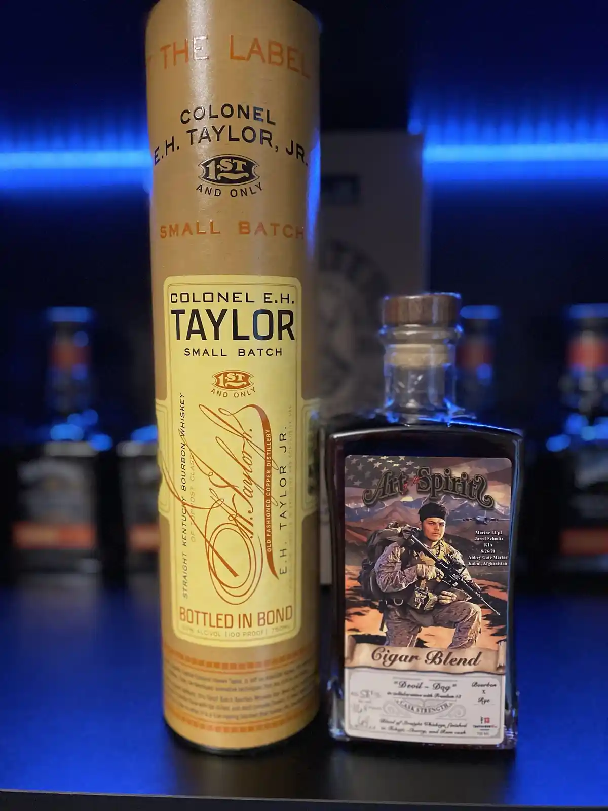 Two whiskey bottles sit side by side on a bar: a tall, cylindrical Colonel E.H. Taylor Jr. Small Batch bottle with a tan…