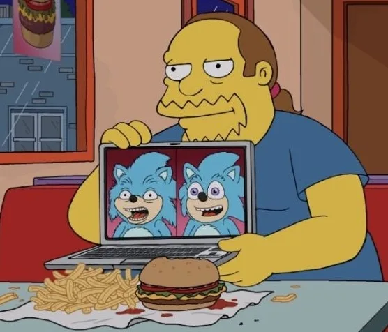 A man with a receding hairline and a blue shirt sits at a table with a messy burger and fries, holding a laptop. The laptop…
