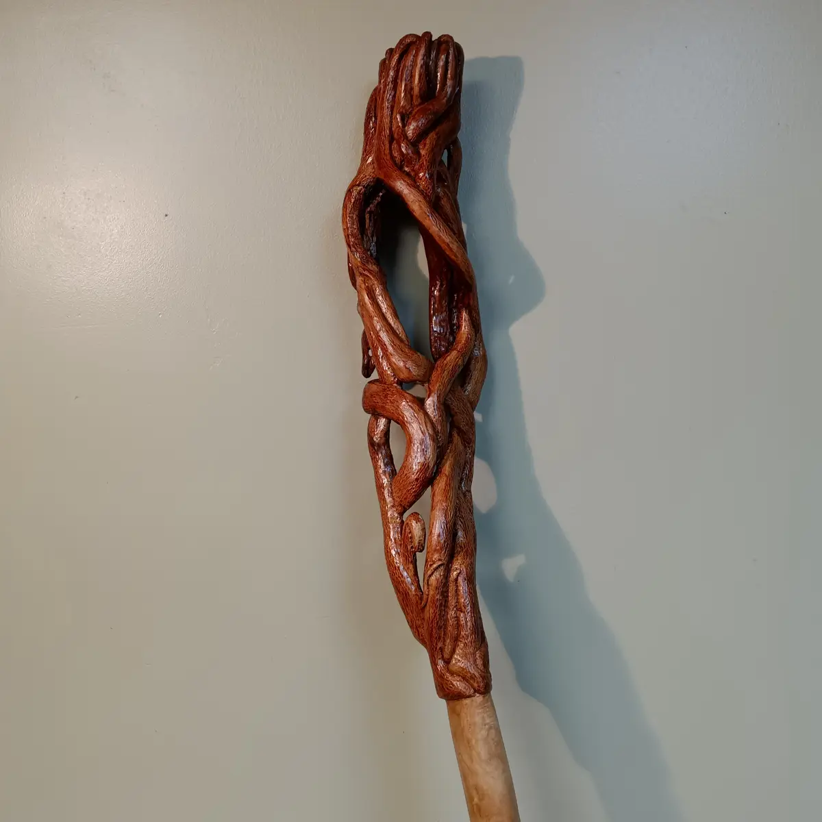 A wooden staff with an ornate, twisted top resembling intertwined tree roots or vines, mounted against a plain wall. The…