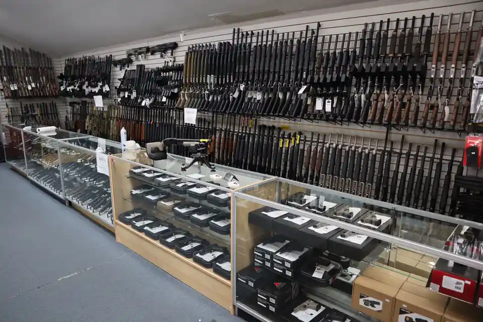 An interior shot of a gun store with walls lined with rifles and shotguns on display racks, and glass cases in the…