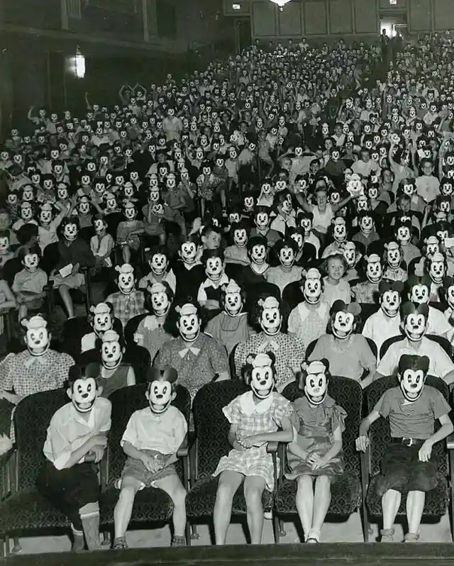 a black-and-white photo shows a large audience seated in a theater, all wearing Mickey Mouse masks, with some raising their…