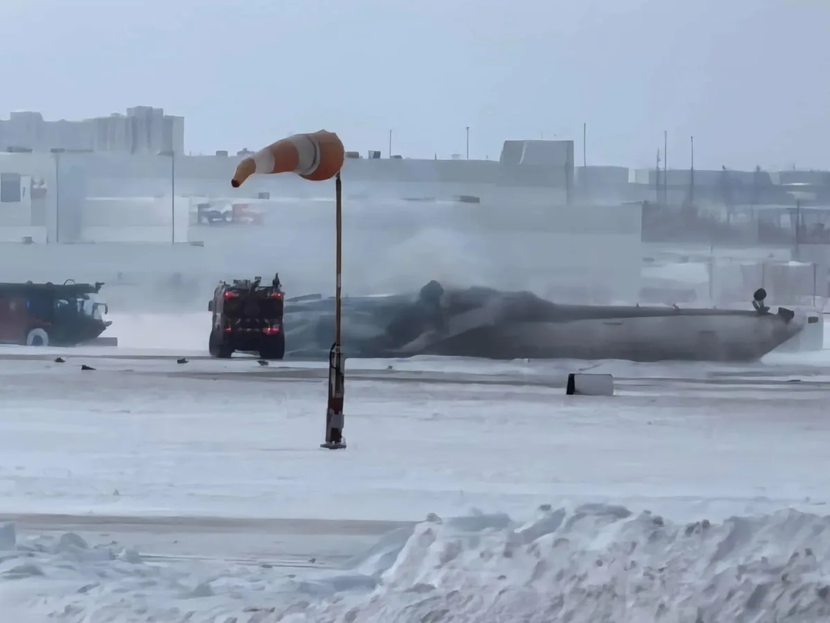 A snowy airport tarmac shows a large orange and white windsock bent sharply to one side, indicating strong winds, while a…