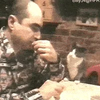 A man sits at a table, initially focused on drinking, then turns his head to watch a black-and-white cat walking behind him…