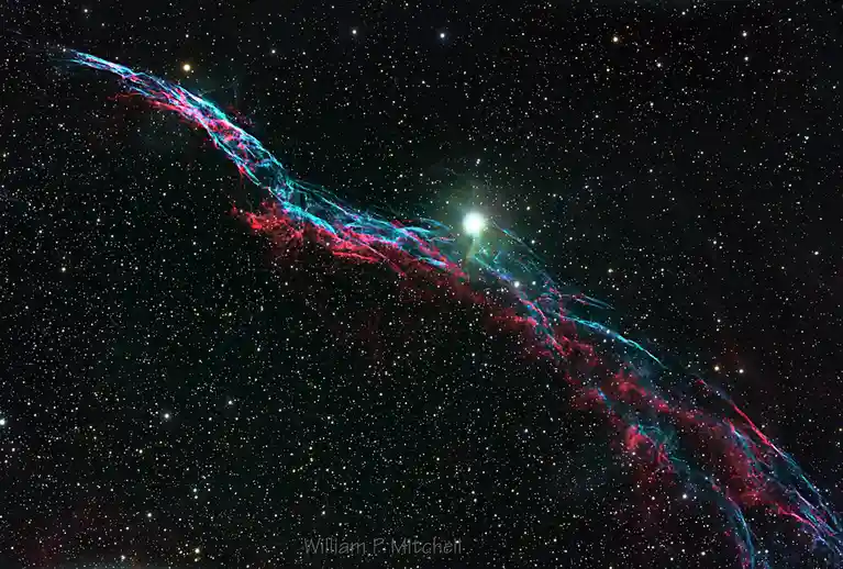 A cosmic image shows a vibrant red and blue nebula stretching diagonally across a star-filled black sky, with a bright…