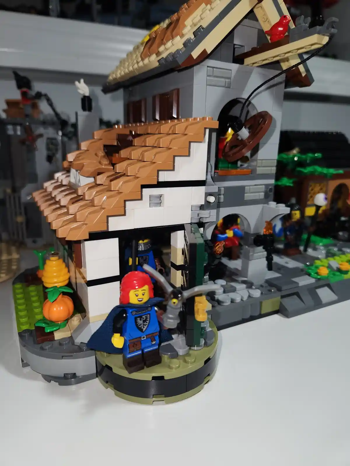 A close-up photo of a LEGO set featuring a detailed building with a brown tiled roof and white walls, a red bird on a beam…