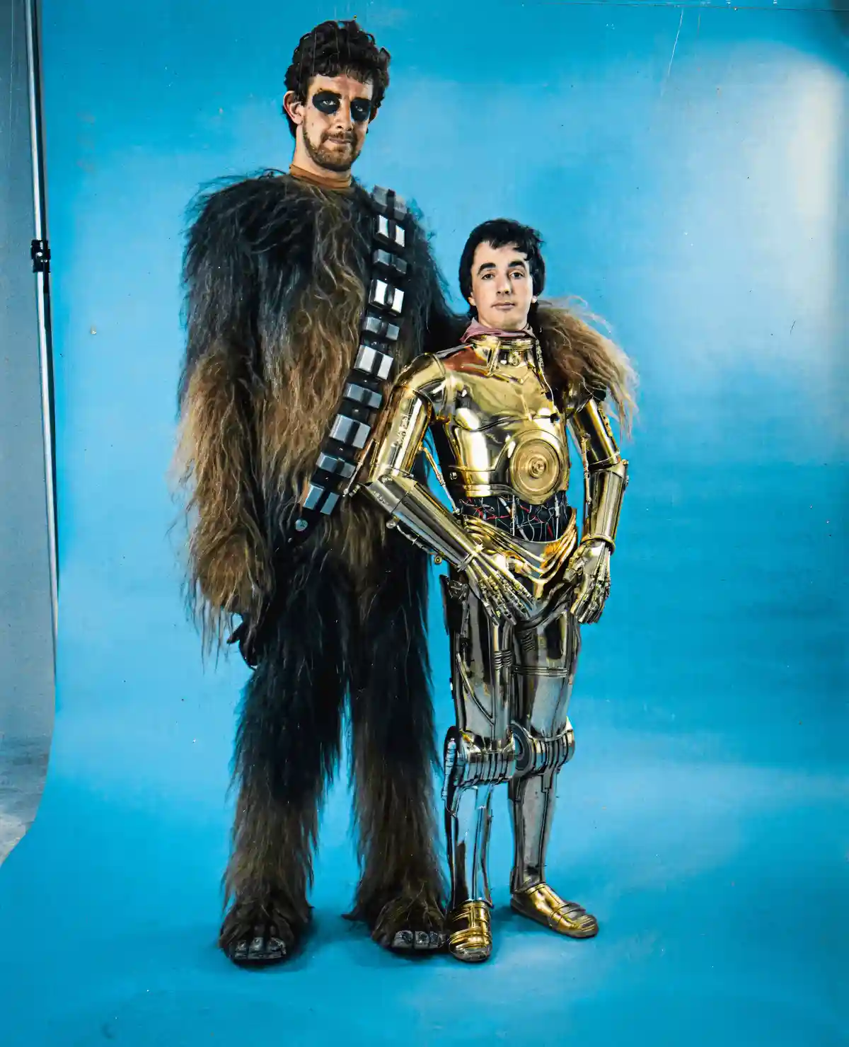 Two men pose against a blue backdrop; one wears a Chewbacca costume with fur and a blaster strap, the other wears a gold and…