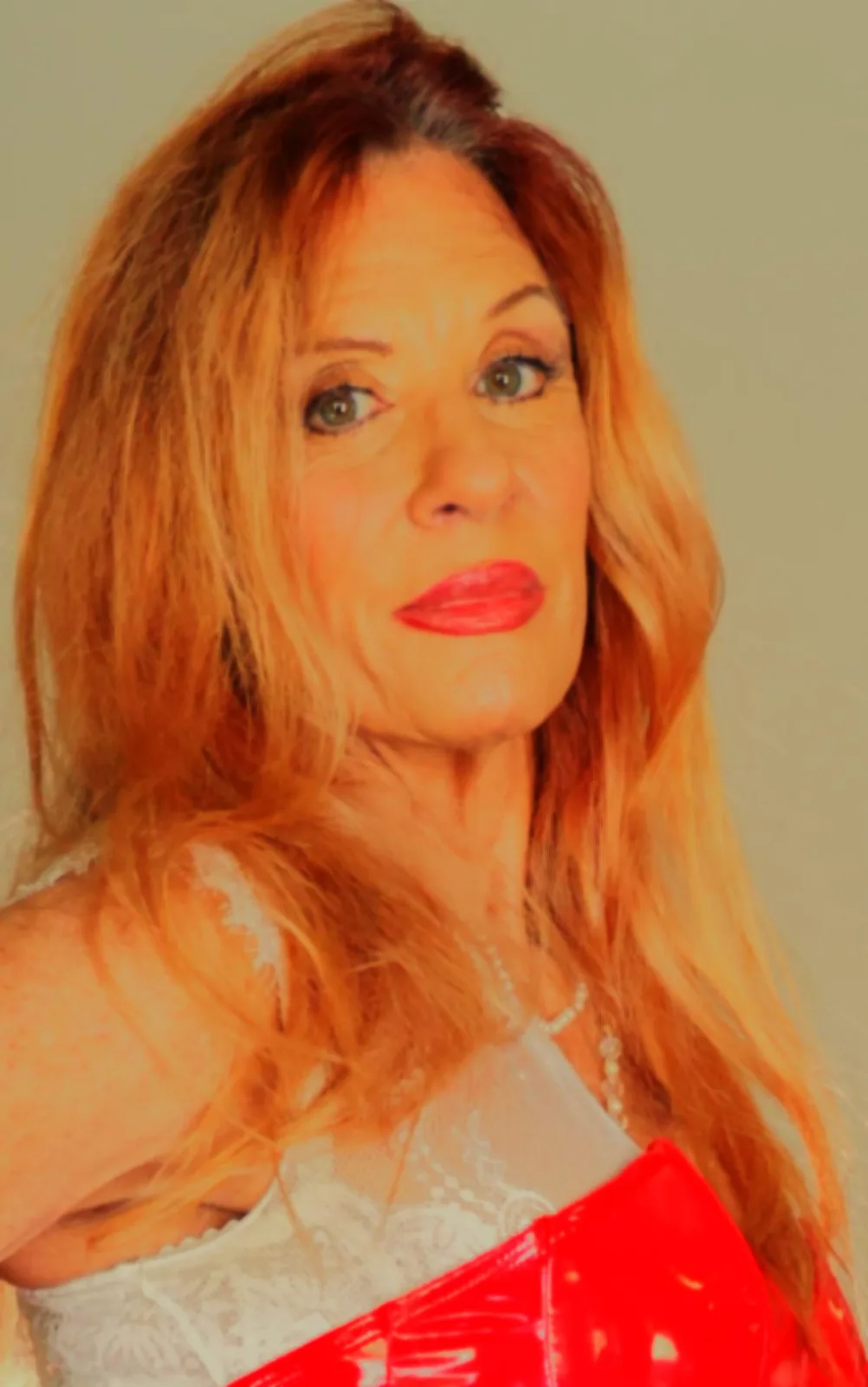 A woman with long reddish-blonde hair, wearing a white lace top and holding a red shiny object, looks directly at the camera…