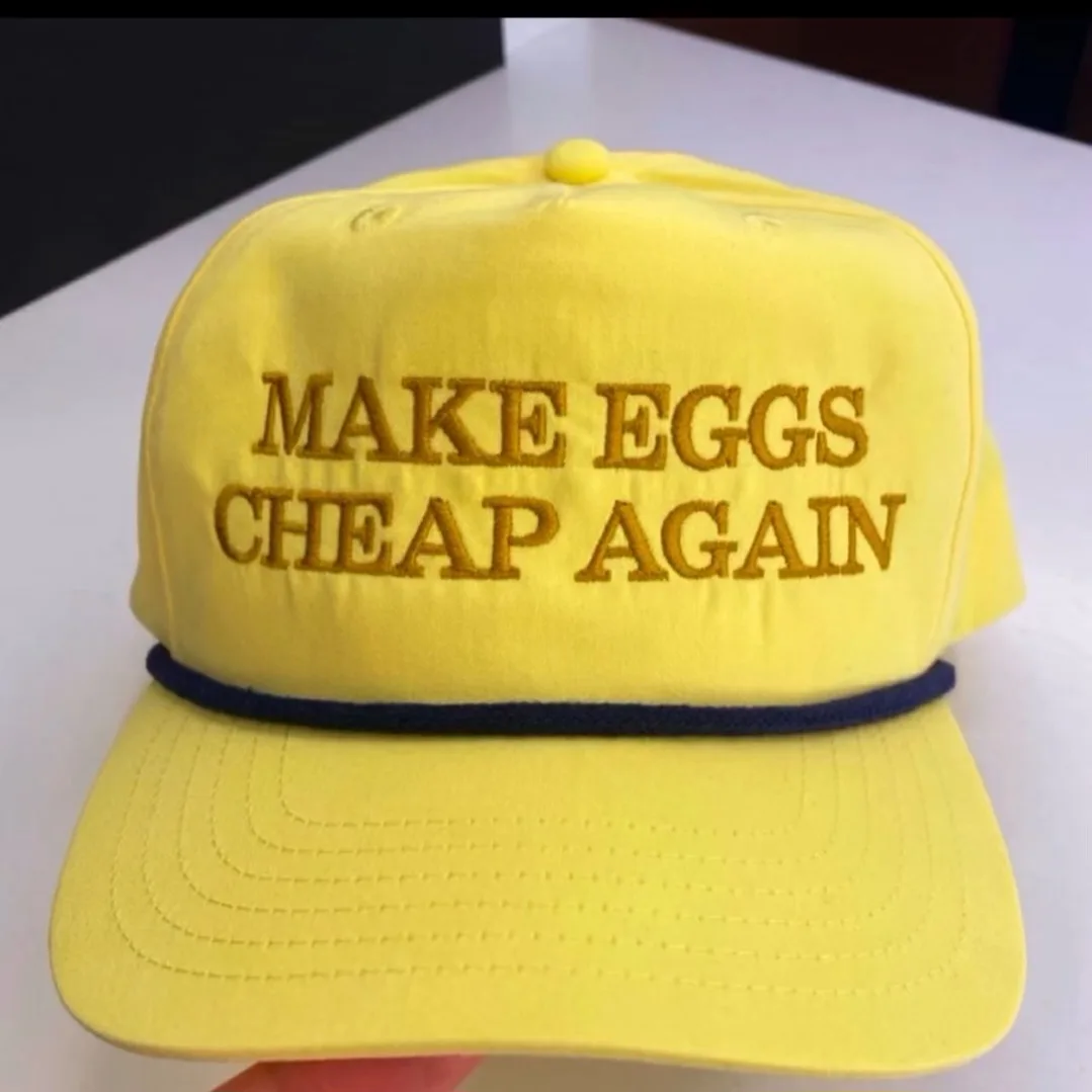 A bright yellow baseball cap with gold embroidered text reading 'MAKE EGGS CHEAP AGAIN' sits on a white surface. The cap has…