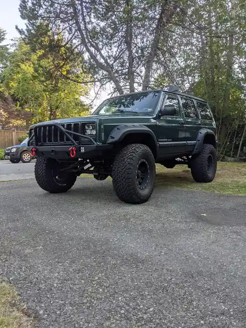 A heavily modified dark green Jeep Cherokee Sport is parked on a residential asphalt driveway, featuring oversized knobby…