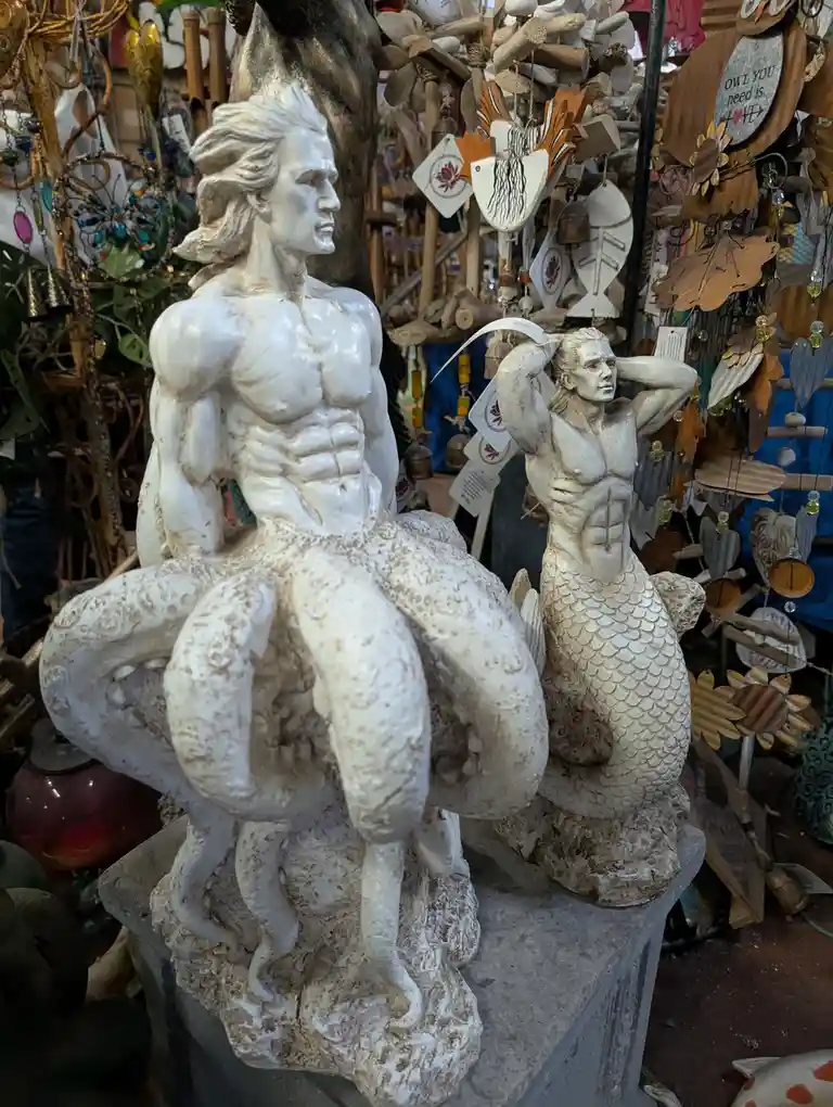 Two white statues of muscular male mermaids sit on a stone pedestal in a craft shop, surrounded by wooden ornaments and wind…