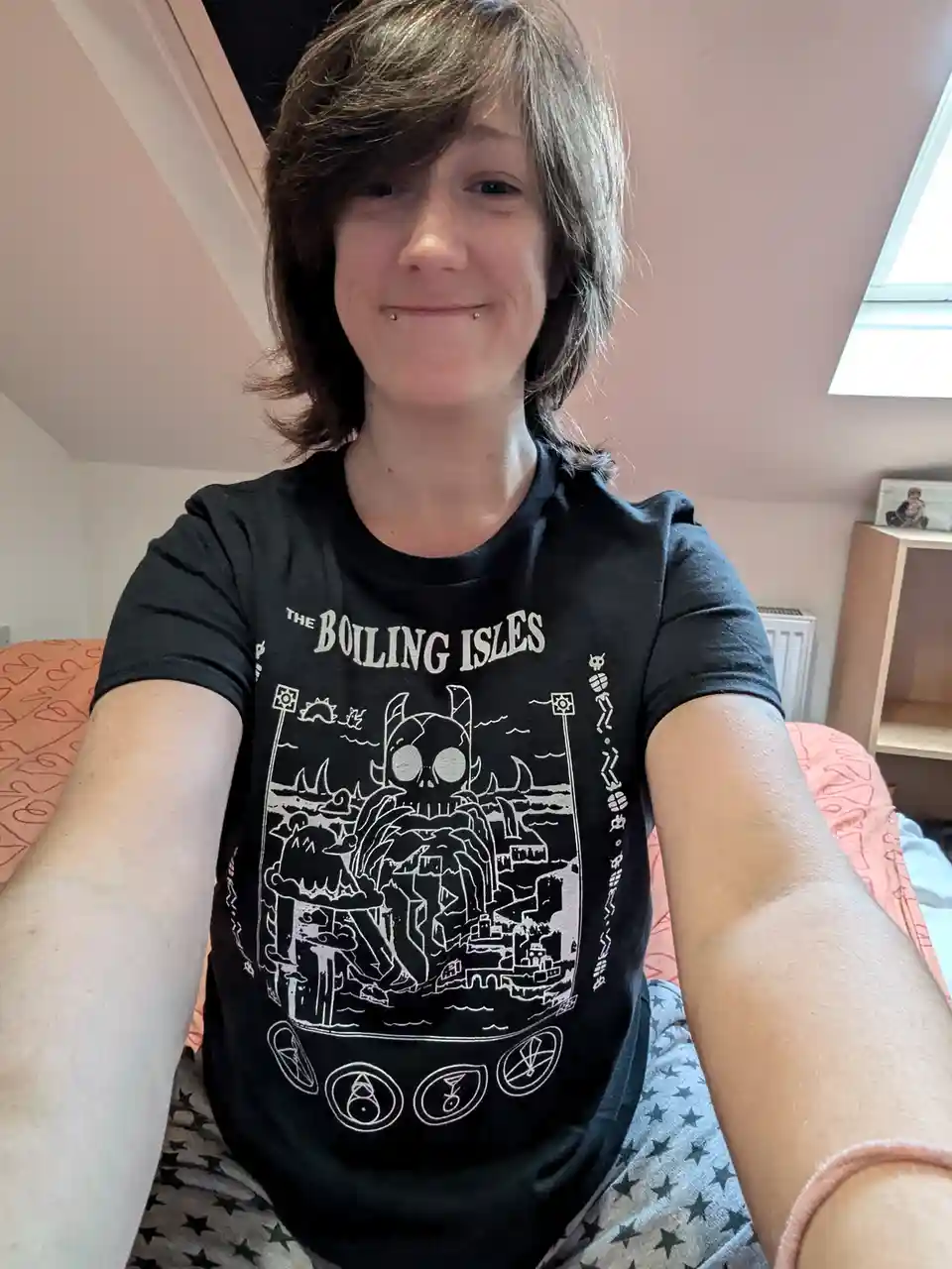 A person with short brown hair and a lip piercing smiles while taking a selfie in a bedroom, wearing a black t-shirt with a…
