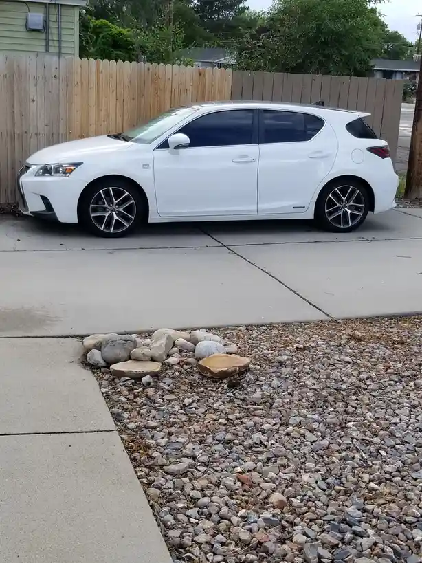 A white Lexus hybrid hatchback is parked on a concrete driveway next to a gravel yard with rocks. Two flat, circular stones…