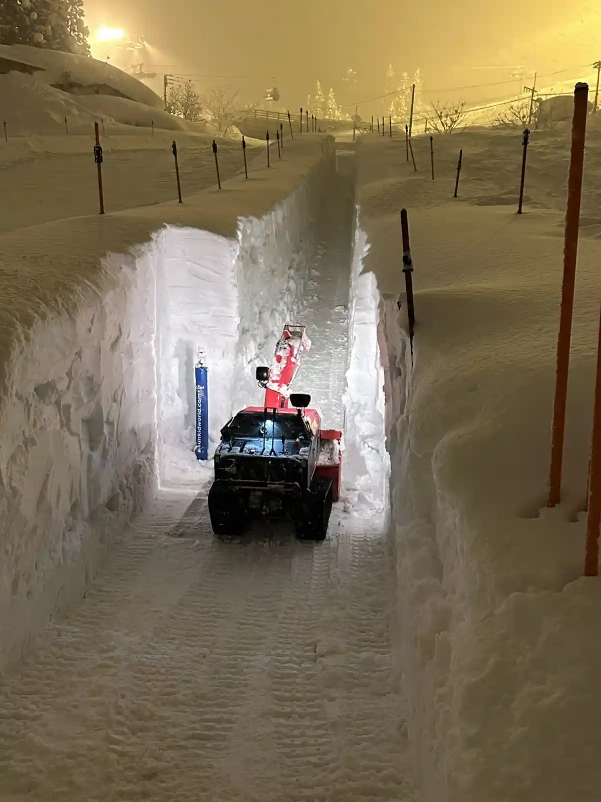 A red and black snowplow vehicle is driving down a narrow, freshly cleared path flanked by towering, sheer walls of snow…