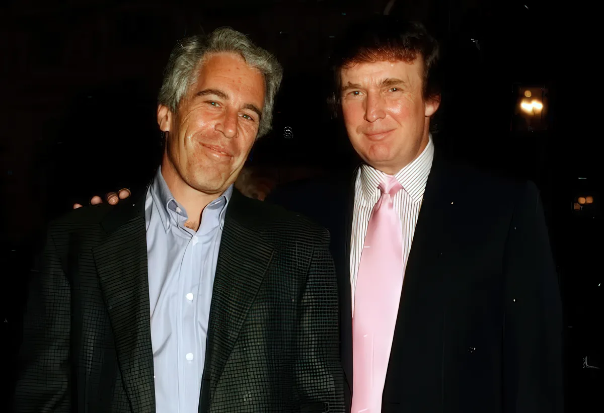 Two men are posing for a photo at a dimly lit event. The man on the left, with gray hair and a patterned jacket, has his arm…
