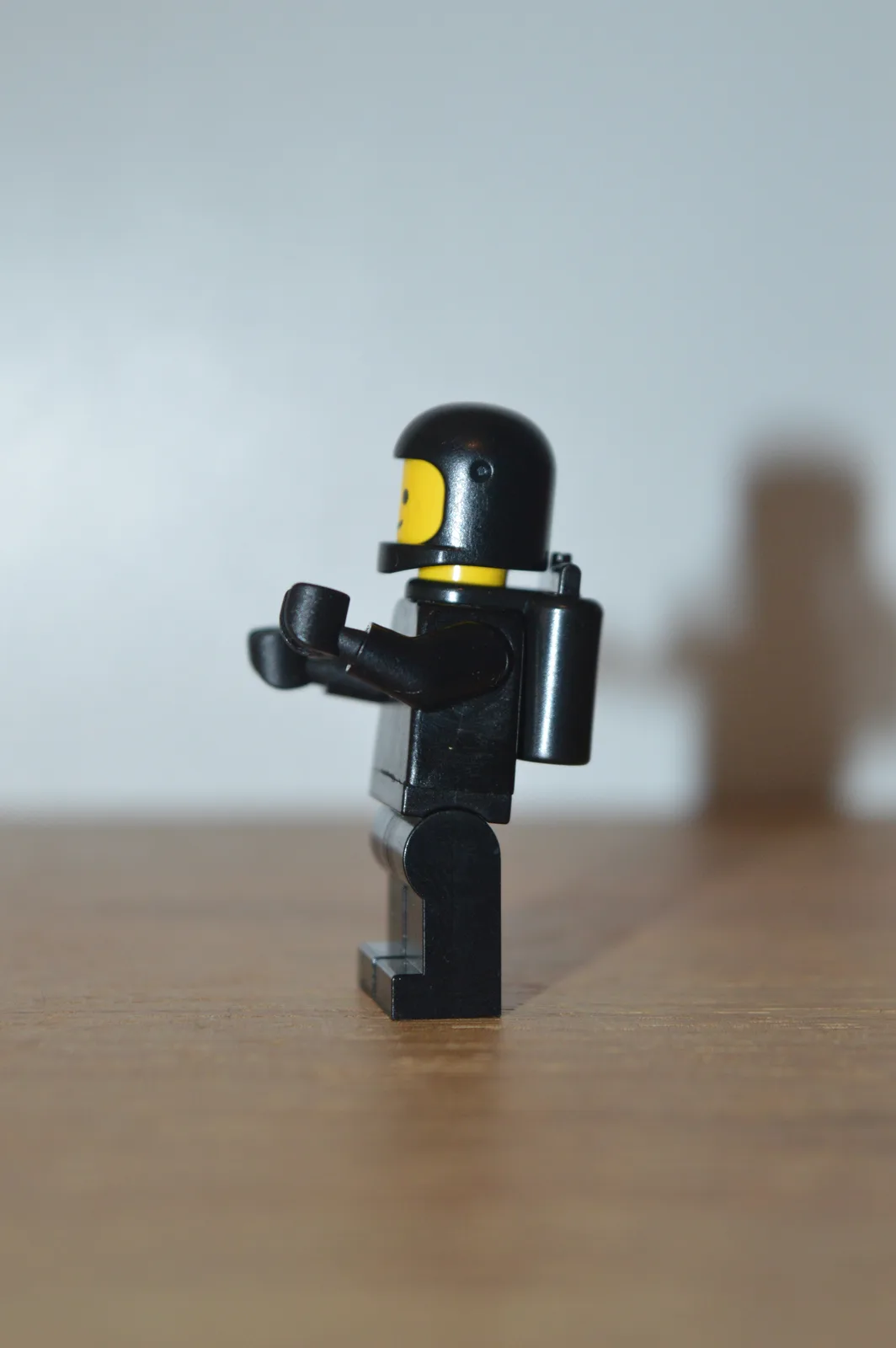 A black Lego minifigure with a helmet and backpack stands on a wooden surface, arms slightly raised. The overlaid text reads…