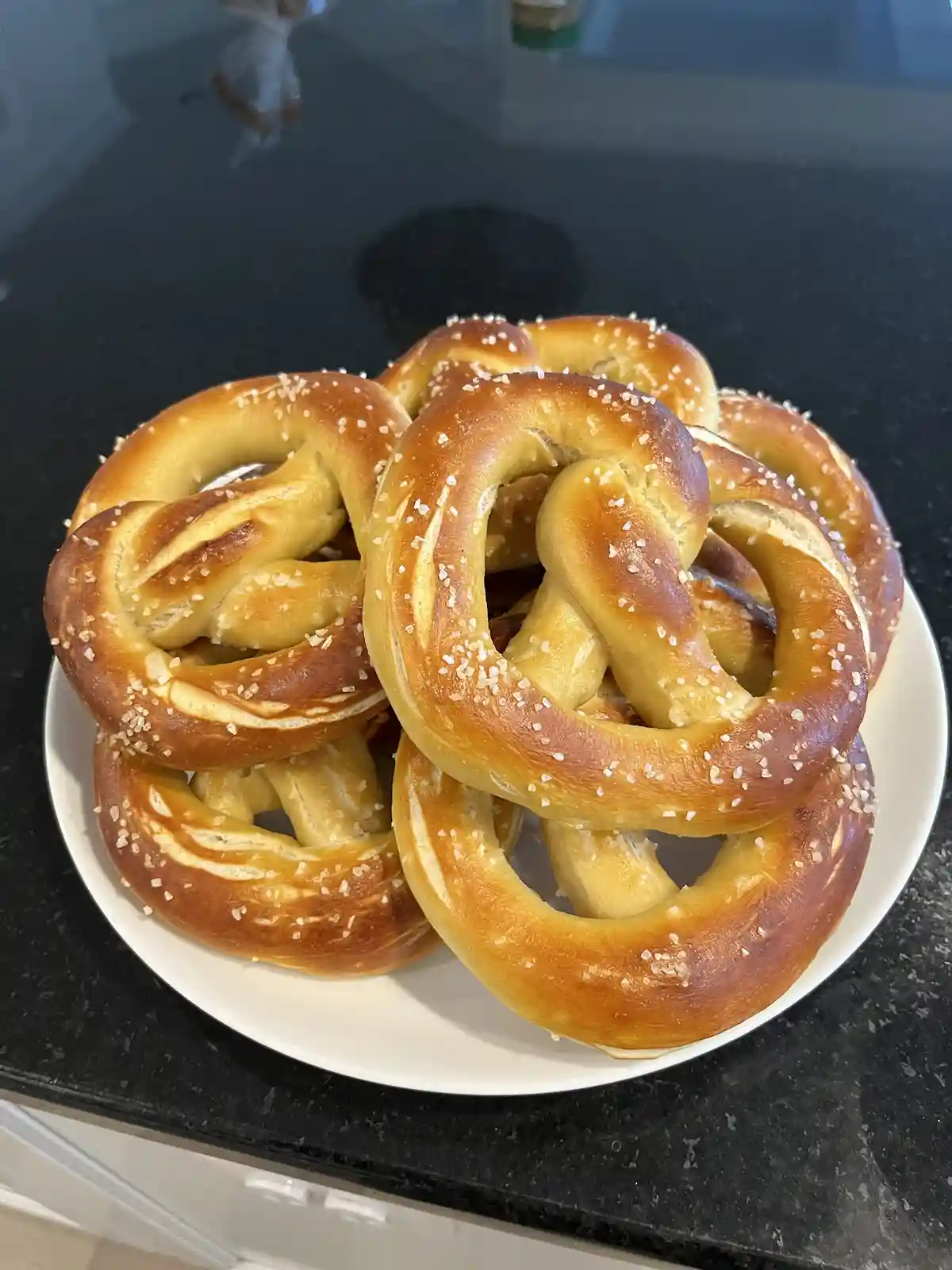 A white plate piled high with freshly baked, golden-brown pretzels topped with coarse salt, sitting on a dark kitchen…