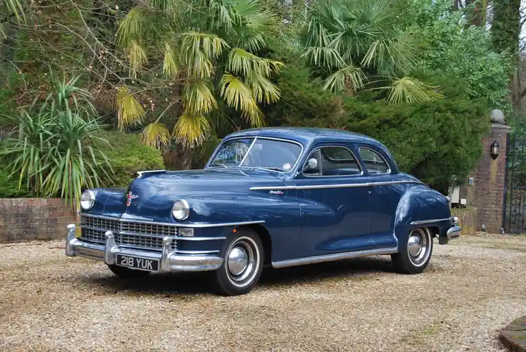 A shiny blue 1940s Chrysler coupe is parked on a gravel driveway in front of lush greenery including palm trees. The car's…