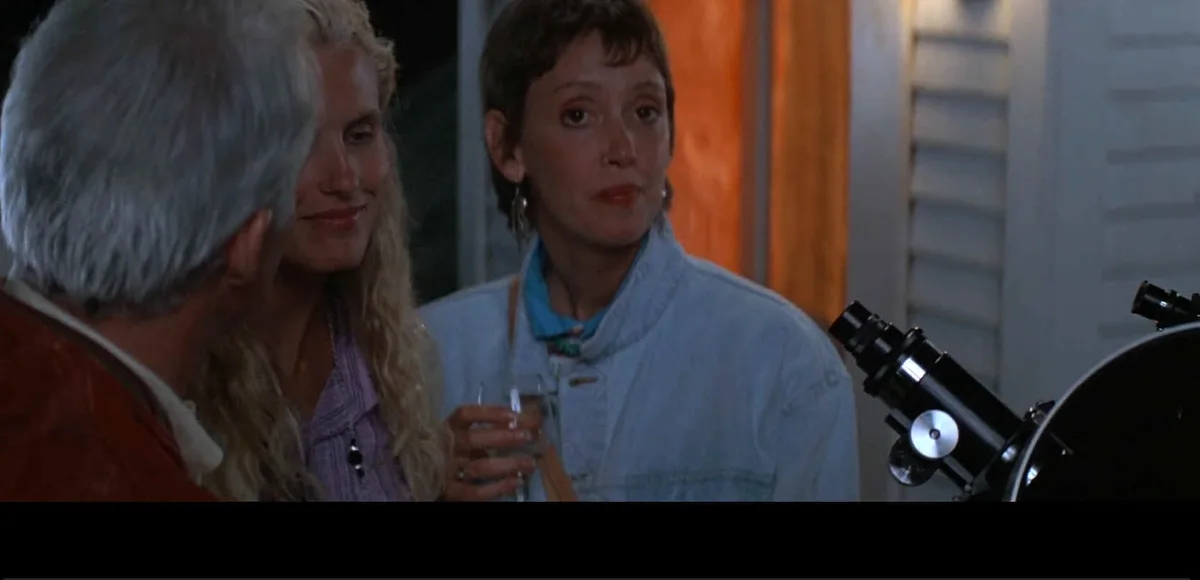 A still from a movie showing three people at an outdoor gathering, one woman in a denim jacket looks confused or unimpressed…