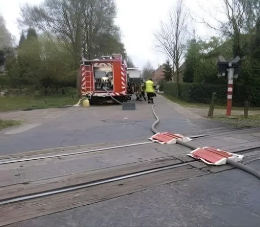 A red fire truck is parked on a road next to train tracks, with firefighters in yellow uniforms working. A long hose snakes…
