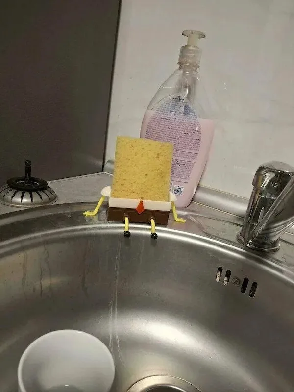 A yellow dish sponge is propped up on a sink ledge, with small yellow pipe cleaners attached as arms and legs, and a tiny…
