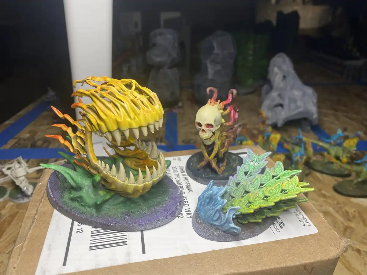 A close-up photo shows three intricately painted fantasy miniatures on a cardboard box. The largest is a monstrous…
