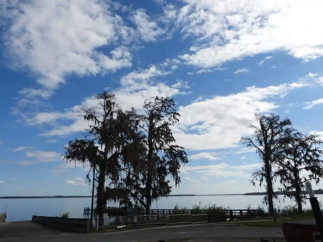 A scenic photo of a calm lake under a partly cloudy blue sky, framed by tall cypress trees draped with Spanish moss. A…