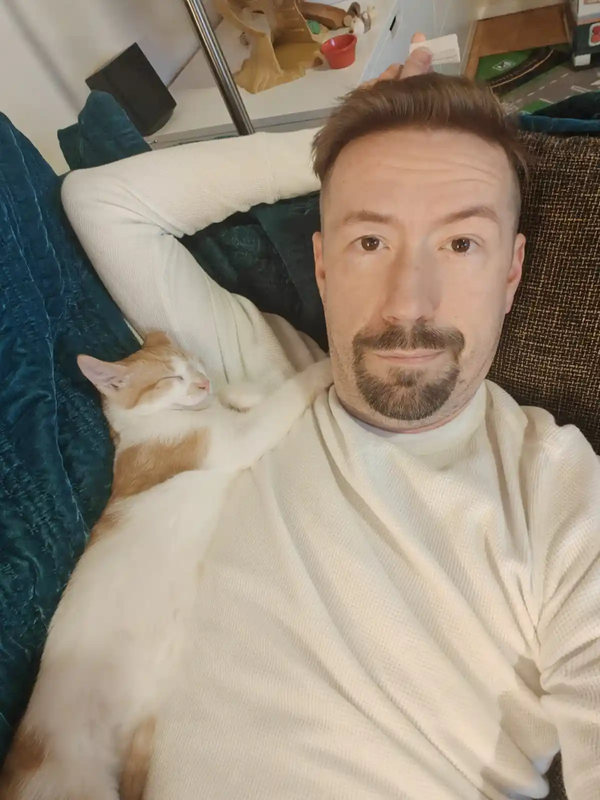 A man in a white sweater lies on a couch with a sleeping orange and white cat draped over his chest and arm. He looks…