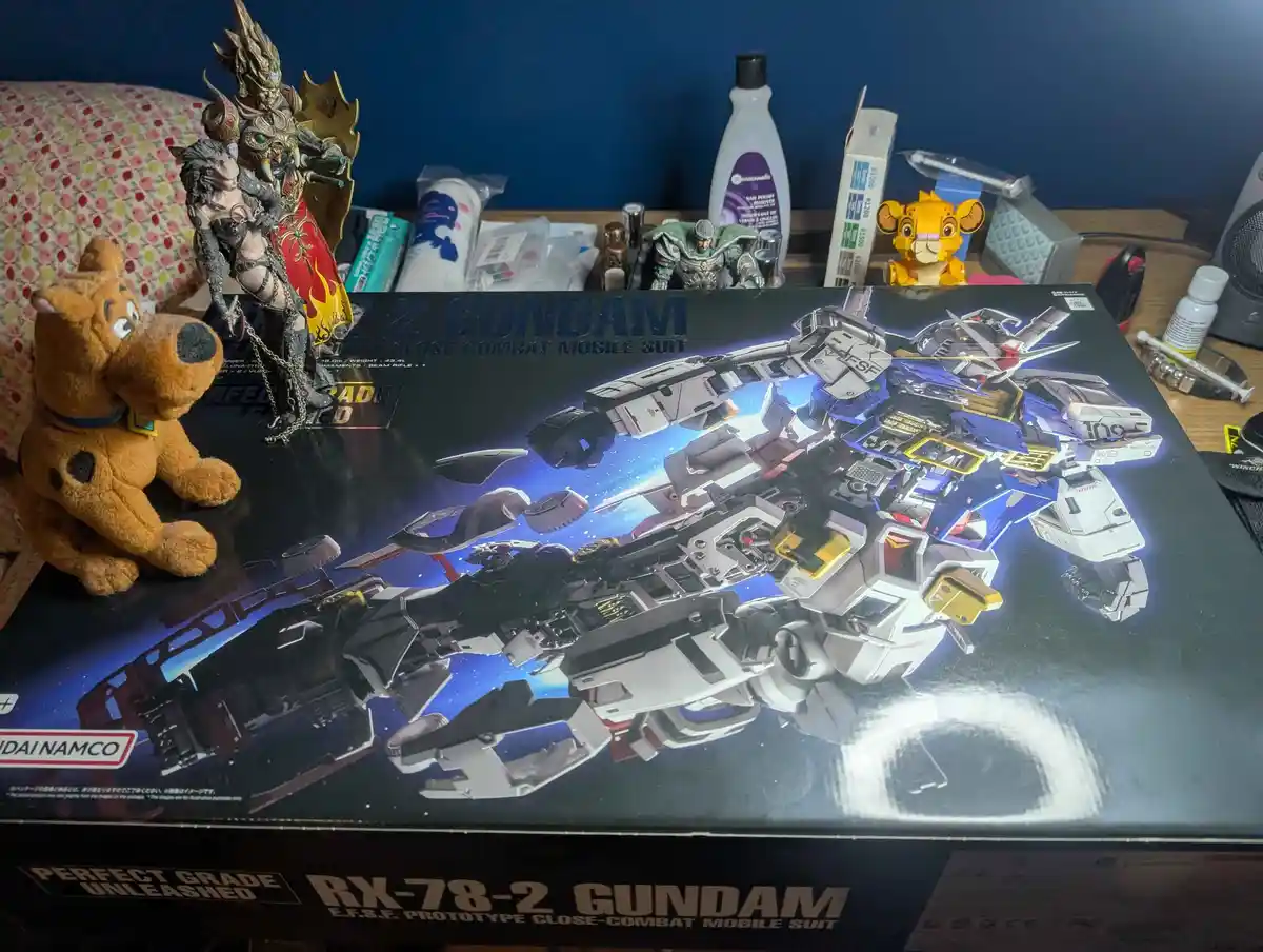 A photo shows a large Gundam model box featuring the RX-78-2 Gundam, with a plush Scooby-Doo toy sitting on the left side of…