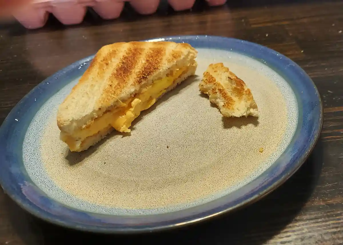 A toasted sandwich cut in half sits on a speckled blue plate, with one half eaten and the other half still intact, revealing…