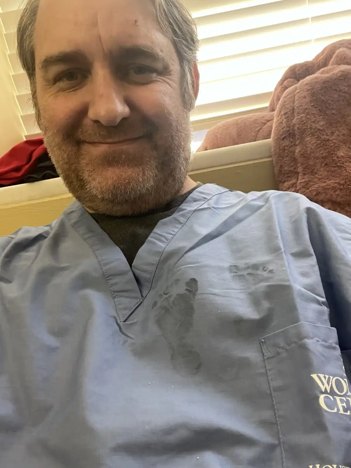 A man in light blue medical scrubs with a visible dark foot print on the front, smiling at the camera while seated on a bed…