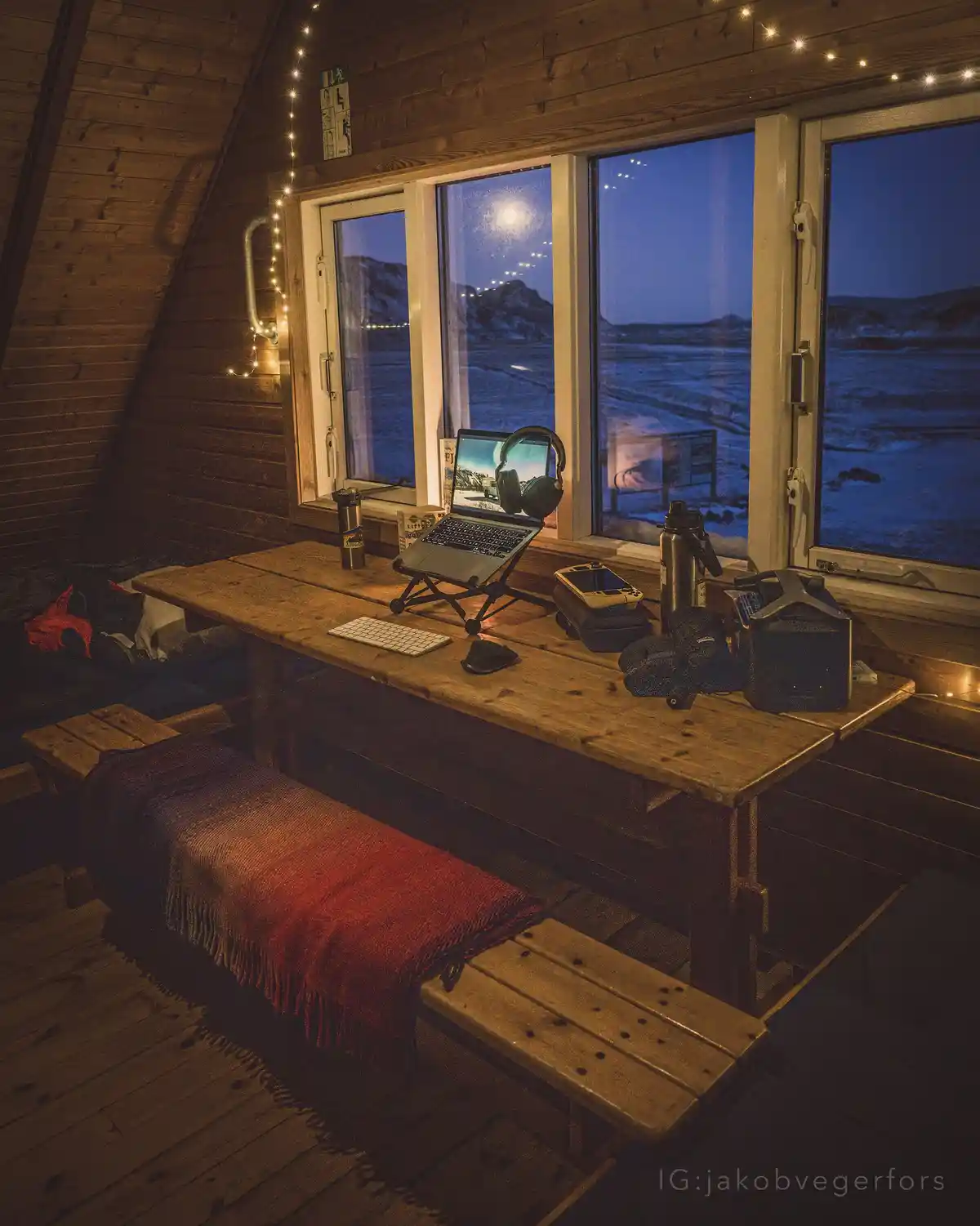 A cozy wooden cabin interior at night, featuring a rustic wooden desk with a laptop, headphones, water bottles, and a red…