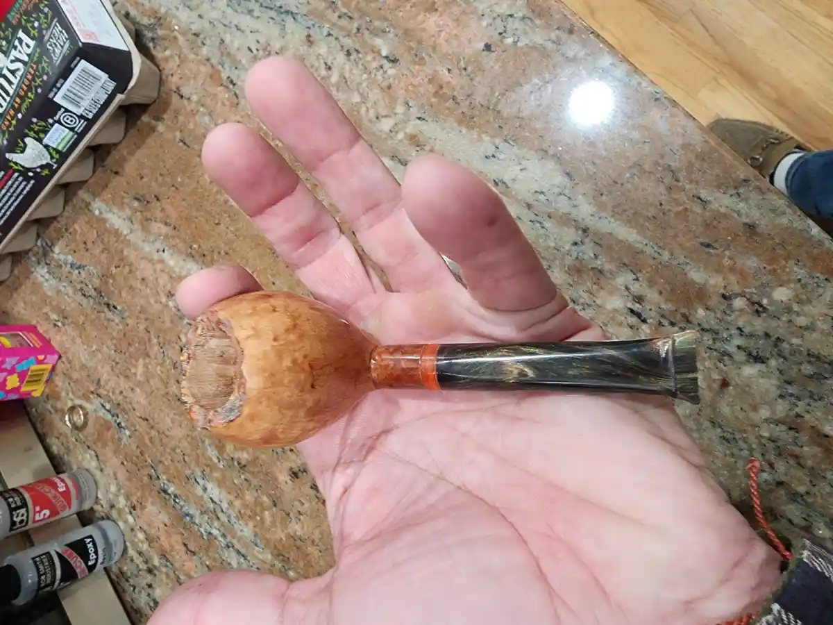 A hand holds a wooden smoking pipe with a visibly repaired, rough-edged bowl, set against a granite countertop with epoxy…