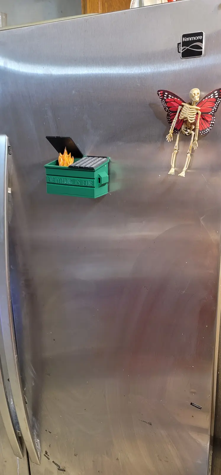 A stainless steel refrigerator door has two magnets: a green dumpster magnet with flames inside and the text 'EVERYTHING IS…